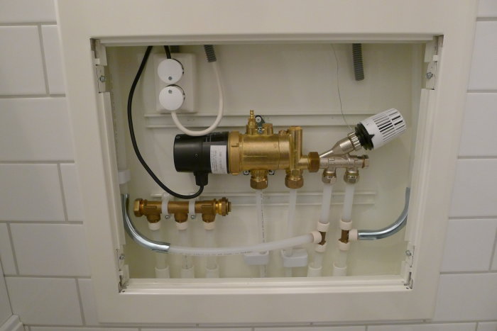 Ett öppet installationsutrymme i en vägg med ett kopplingsschema för golvvärme, inklusive rör, ventiler och en ställdon.