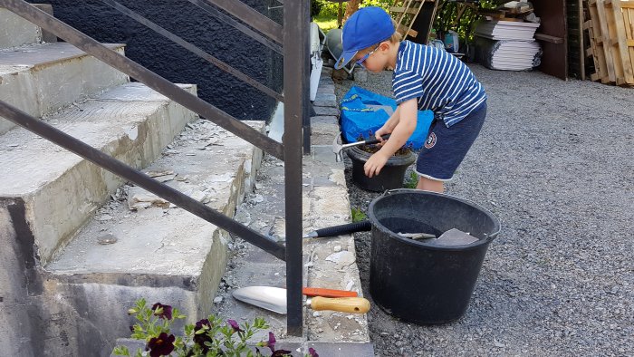 Barn i keps plockar upp bitar av klinker vid en renoverad trappa med byggverktyg i förgrunden.