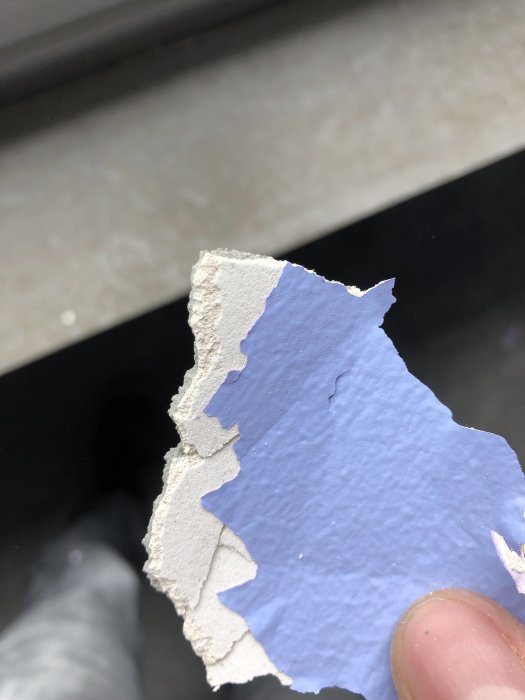 Närbild på hand som håller tapetbit med lager av färg, spackel och tapetklister som avlägsnats från vägg.