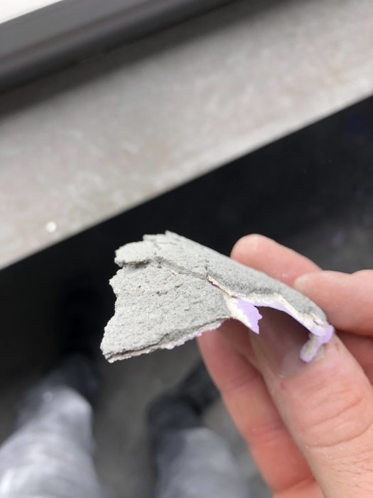 Närbild av en hand som håller en skrapad väggbit som visar lager av spackel, färg och tapet.