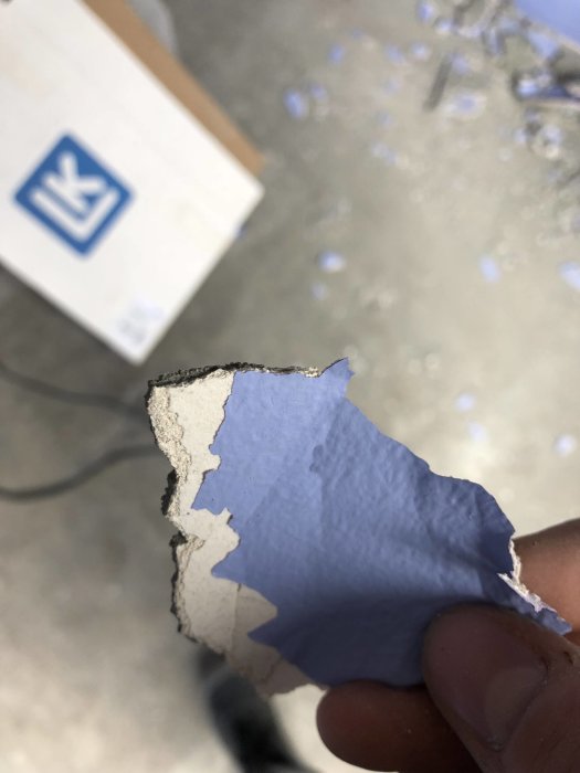 Närbild på en hand som håller en bit borttagen blå tapet med underliggande putslager.