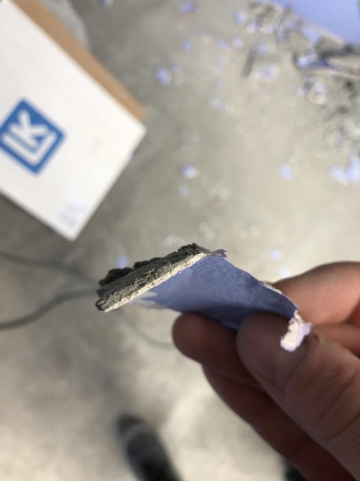 En hand håller en bit av väggbeklädnad med flera lager – betong, puts, spackel, färg och tapet.