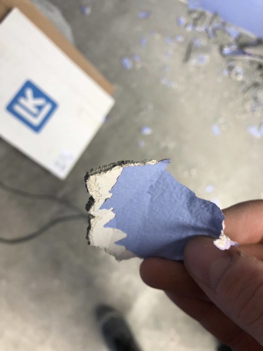 Hand håller ett bit tapet med flerskikt av färg, spackel och tapetklister som skalats av en betongvägg.