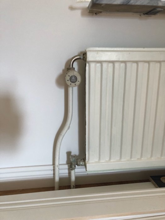 Vit radiator med termostatventil och ovanligt placerade rör som kommer upp från golvet till vänster om fönstret.