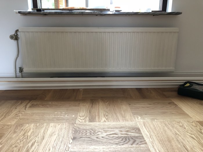Radiator framför fönster med synliga rör som leder upp från golvet till vänster i bilden.