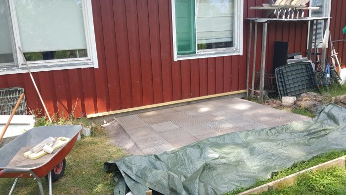 Nylagd plattgång framför röd trähusvägg med en skottkärra och byggmaterial åt sidan.