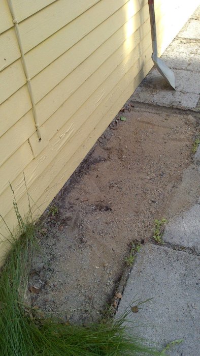 Hörn av hus med borttagna stenplattor som avslöjar fin sandig mark intill en gul vägg och gräs.