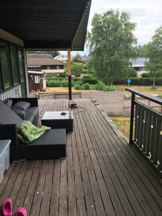 Utemöbler på en träaltan med överbyggd del, utsikt mot trädgård och bostadshus, rosa skor i förgrunden.