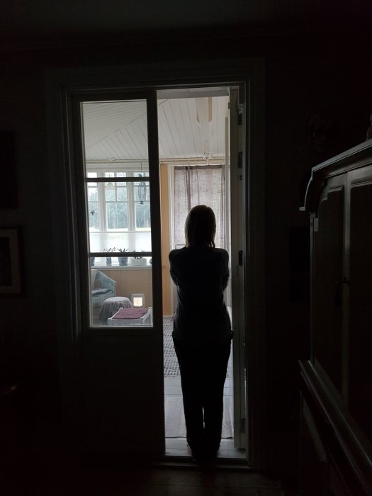 Kvinna står i en dörröppning och tittar ut på ett ljust rum, kontur mot ljus bakgrund.