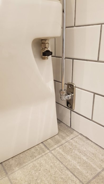 Vit toalett med anslutning i metall och manchett till vägg, fläns felplacerad, kakelvägg och klinkergolv i bakgrunden.
