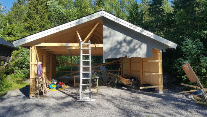 Pågående byggnadsarbete av träbyggnad med ny panel på framsidan, trädgårdsverktyg och byggmaterial runt omkring.