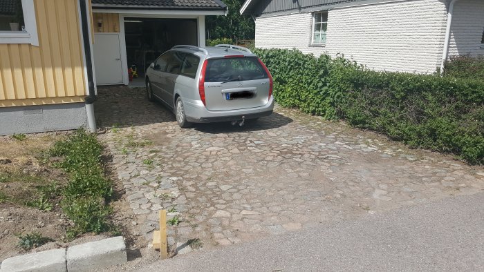 Bil parkerad på sliten stenbelagd uppfart vid hus med öppen garagedörr och grönska.