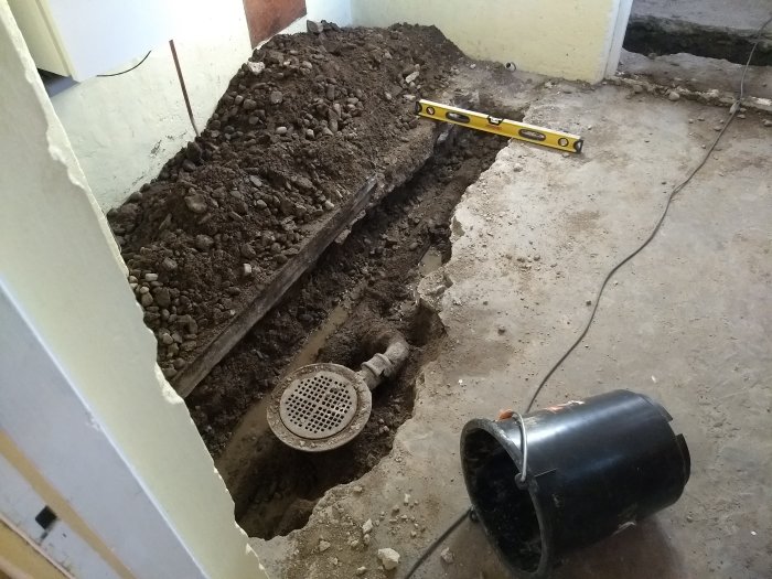 Uppbrutet golv i ett rum med synlig avloppsbrunn, omgiven av jord och grus, med ett vattenpass och hink i förgrunden.
