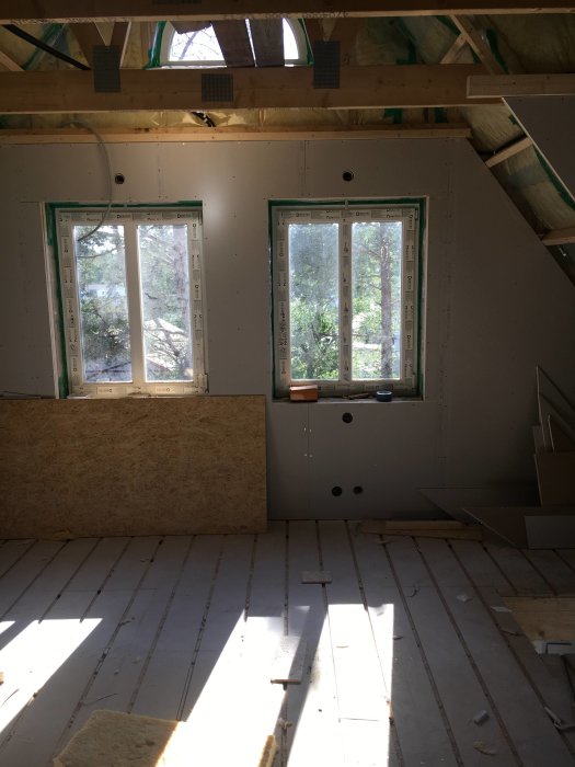 Inre del av ett rum under renovering med oisolerade väggar och fönster samt trägolv och takstolar.