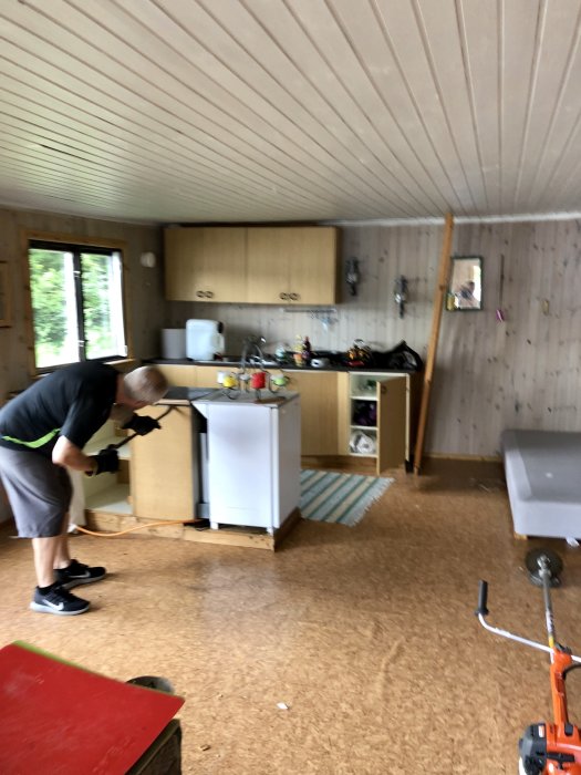 Person i arbetskläder bär verktyg och börjat montera ned ett kök, tecken på renovering pågår.