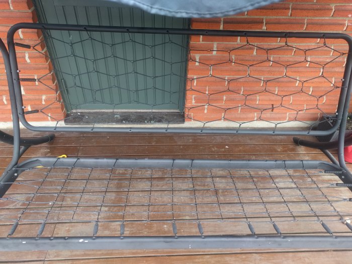 Svart hammockställning med ett slitet nät utan flera krokar framför en tegelmur och fönster.