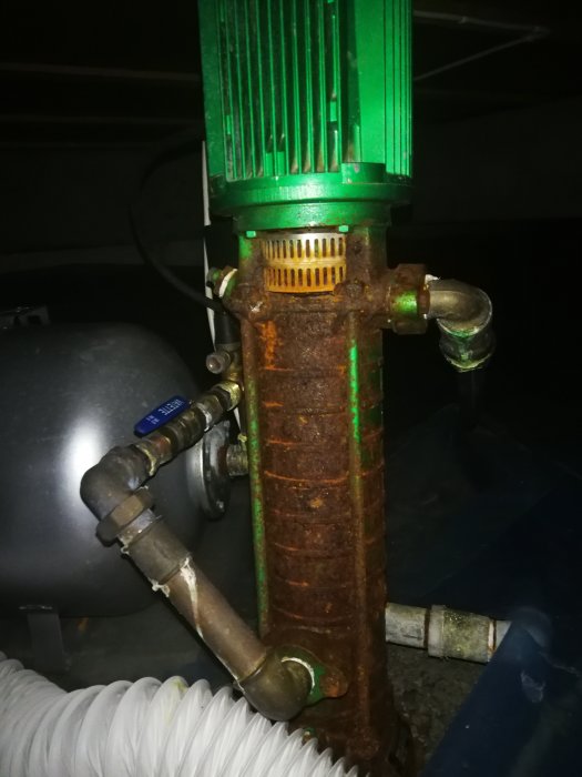 Rostig hydroforpump kopplad till vattenrör och en blå hydrofortank i ett mörkt utrymme.