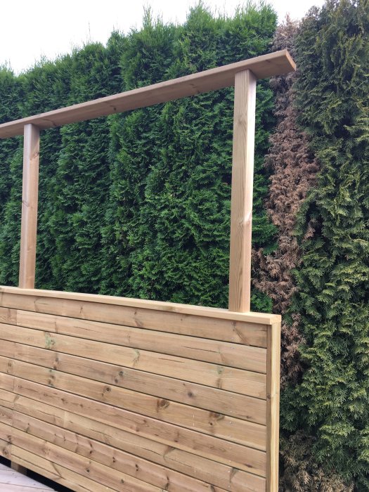 Trädäck med överliggare på vertikala stolpar framför en häck av gröna och bruna buskar.