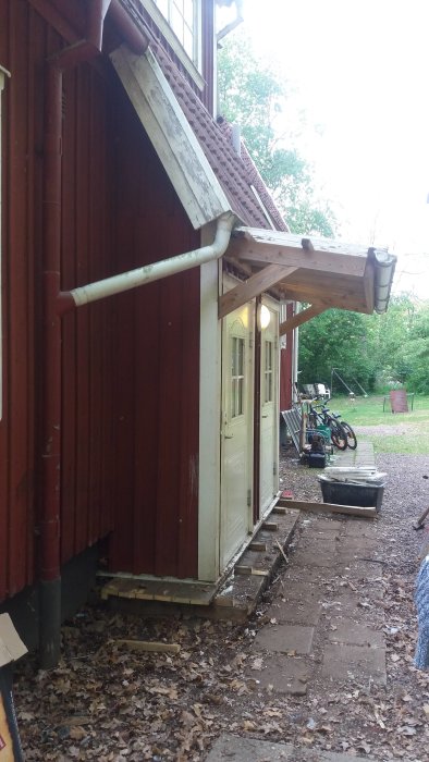 Rödfärgat hus med löst hängande stuprör vid takfot nära huvudentré och nedblåsta löv på marken.