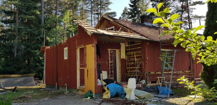 Hus under renovering med takstolar delvis nedtagna och väggar i rivningsprocess, omgiven av grönska.