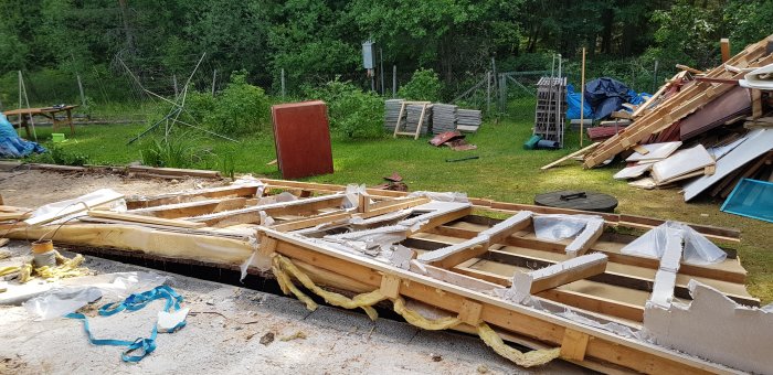 Nedrivna takstolar och spikrester på en byggplats med spridda byggmaterial i bakgrunden.