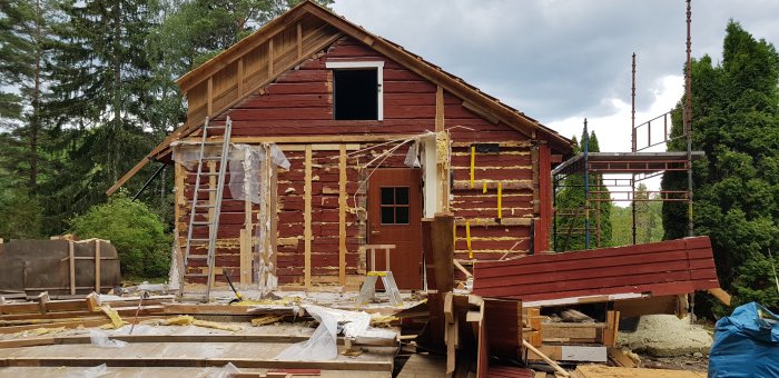 Hus under renovering med borttagna väggar och takstolar, omgivet av byggavfall och en skogsbackgrund.