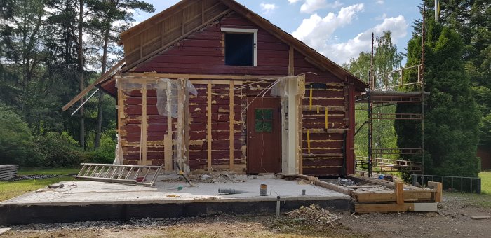 Rödfärgad stuga under renovering med borttagna väggsektioner och isolering synlig, omgiven av byggnadsställning.