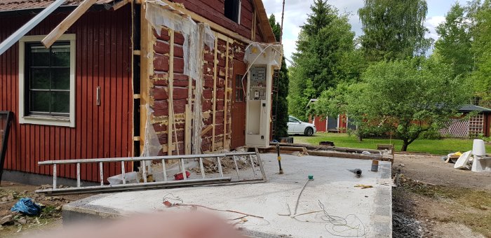 Renoveringsarbete på ett rött trähus med väggar delvis rivna, ställning på marken, och skog i bakgrunden.