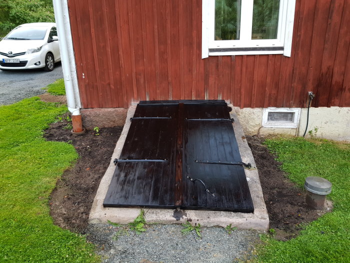 Nya svarta luckor installerade på källaringång bredvid röd trävägg och vit bil.