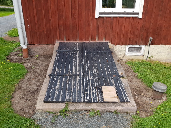 Gamla slitna källarluckor av trä ligger på betongplatta vid rödmålad husvägg.