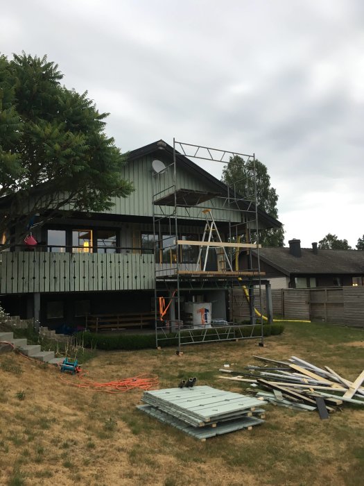 Omonterad stålbyggnadsställning framför hus med byggmaterial på gräsmatta.