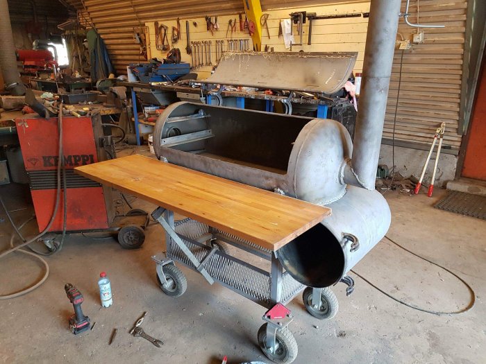 Hemmagjord mobil arbetsbänk med en träskiva monterad på en gammal cylindrisk metalltank på hjul i en verkstad.
