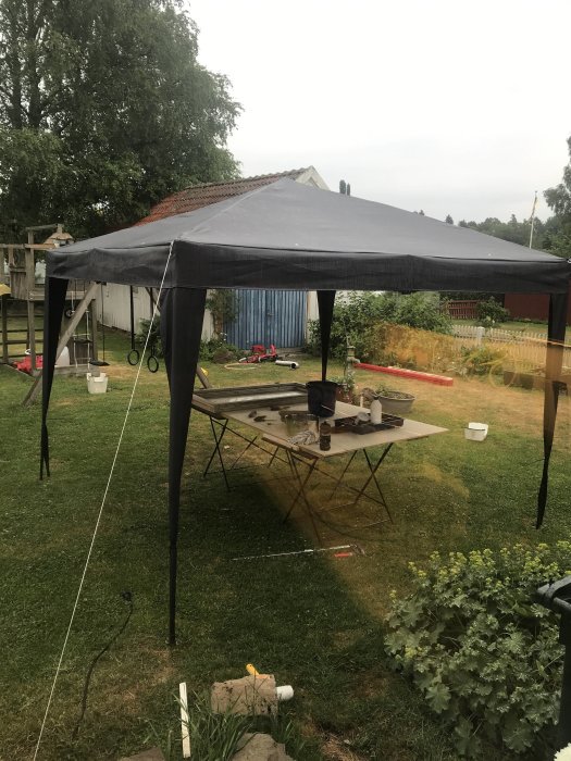 Partytält som används vid fönsterrenovering med verktygsbord, omgivet av trädgård och hus.