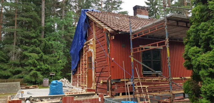 Röd trästuga under renovering, med borttagen utbyggnad och presenning över taket, omgiven av skog.