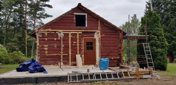 Rödmålad timmerstuga under renovering med borttagen panel och skydd med presenning, omgiven av byggmaterial.