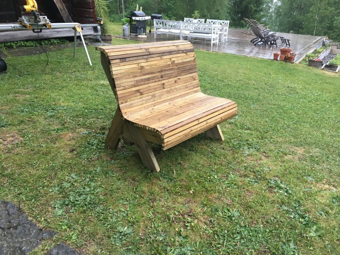 Hembyggd träbänk på gräs med våt trädäck och trädgårdsmöbler i bakgrunden.