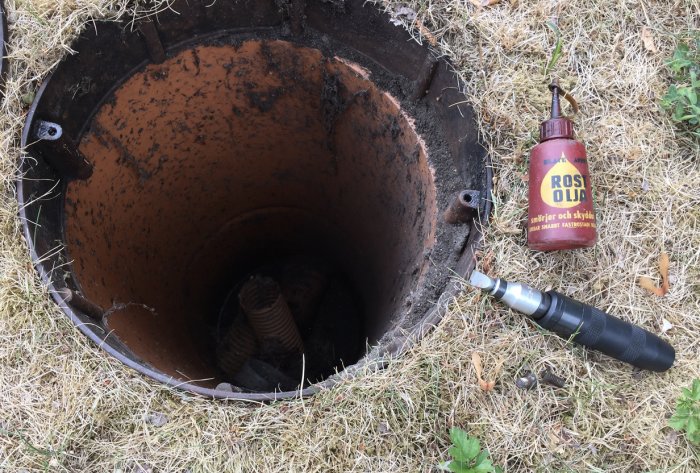 Öppet brunnslock med synliga spår av rost, en flaska rostolja och en 12 mm slagskruvmejsel på gräsytan.