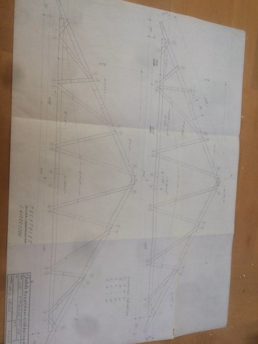 Svårlästa, upprätthållna arkitektoniska ritningar av takstolar med märkningar och mått, placerade på ett träbord.