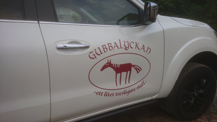 Vit bil med reklamdekor "GUBBLYCKAN -ett litet trevligare stall-" och bild av en stiliserad häst.