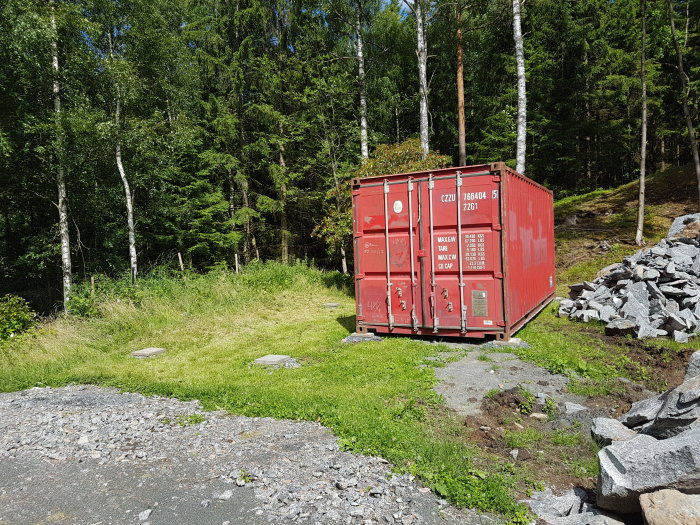 Röd 20 fots fraktcontainer på betongplattor i en trädgård med skog i bakgrunden och en stenhög till höger.