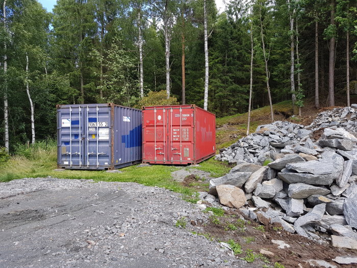 Två fraktcontainrar på en grusplan med skogsbakgrund och en stenhög.