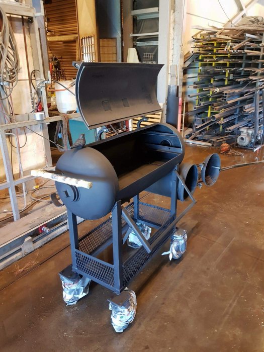 En nybyggd svart smoker grill på hjul i en verkstad, delvis täckt med skyddspapper.