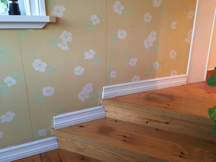 Trappa i ett hem med två trästeg leder upp till en vägg med blommigt tapetmönster, indikation på hemrenoveringsprojekt.