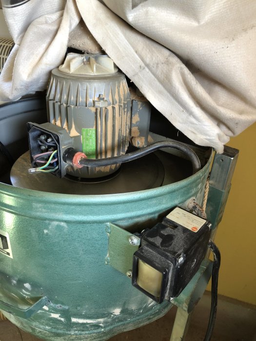 Detaljbild av en grön elektromotor med anslutna kablar och påmonterad svart strömbrytare, delvis täckt av ett tyg.