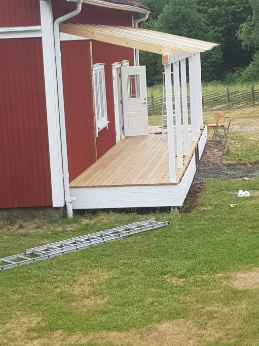 Pågående bygge av trätrapp på en ny veranda med vita stolpar och röd husfasad i bakgrunden.