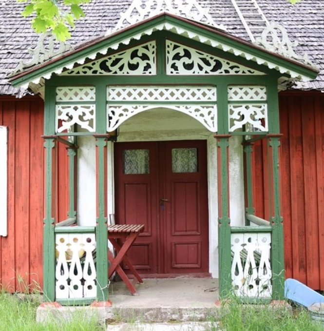 Röd trästuga med detaljerat grönt och vitt verandaräcke, röd dörr och hopfällbar stol.