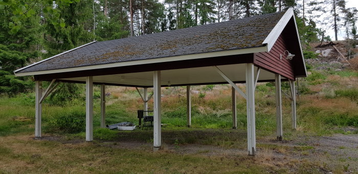 Öppet carportliknande förråd med två fack, oisolerat tak belagt med mossa och vita stödpelare i grön naturmiljö.