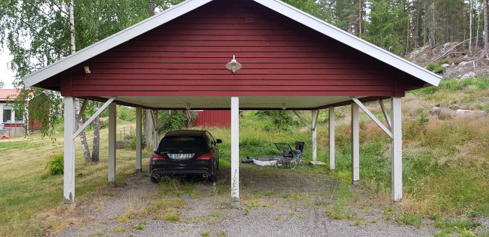 Rödmålat öppet carporttak med en bil parkerad och en släpvagn till höger, planerat för att vara förråd och verkstad.