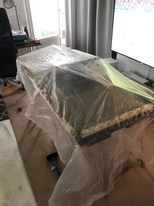 En bord dækket med plast i et rum, klar til at vande hver 24 timer. Arbejdsområde set ved siden af en tændt tv-skærm.