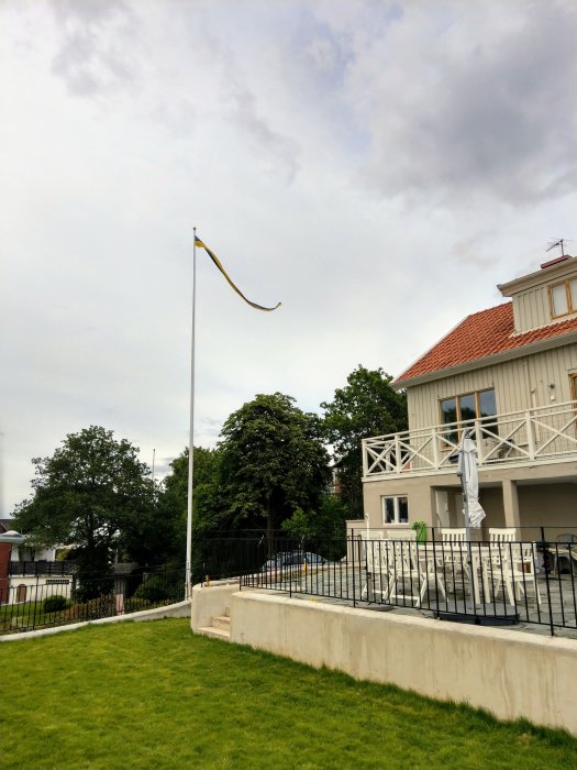 En gul och blå flagga vajar på en gammal flaggstång framför en vit villa med grönt gräs i förgrunden.